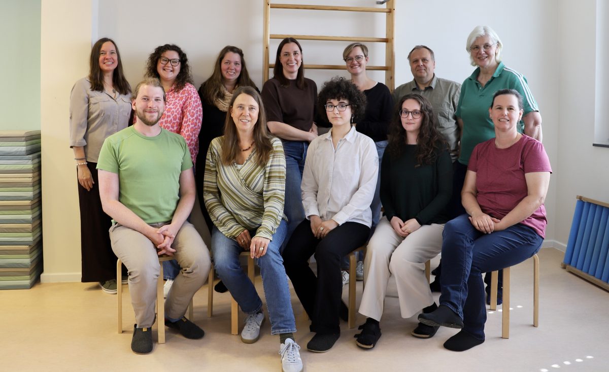 Gruppenfoto des Praxisteams: zwölf Teammitglieder sitzen und stehen lächelnd in einem hellen Therapieraum vor einer Sprossenwand.